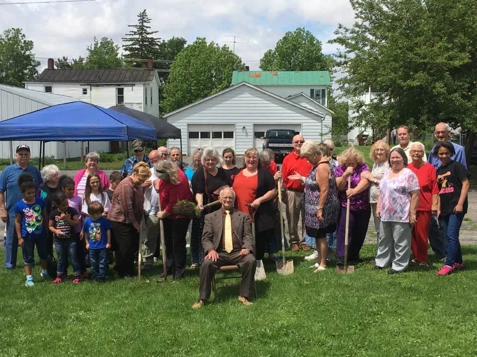 The church family gathered on a lawn with shovels and a piece of turned sod, an older man in a suit seated in front